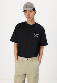 Camiseta de algodón negra con logo en cursiva blanca en el pecho izquierdo, combinada con pantalones cargo de color beige claro, modelo que lleva una gorra gris clara.