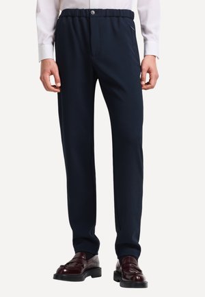 Uomo in pantaloni eleganti blu navy, camicia bianca a maniche lunghe e scarpe eleganti in pelle bordeaux scuro, in piedi contro uno sfondo neutro.
