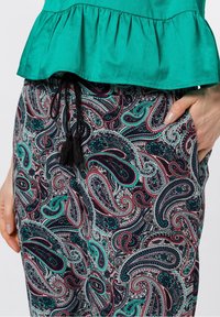 Haut peplum teal associé à un pantalon à motifs avec des designs paisley en rouge, vert et bleu, avec des détails en cordon noir et des glands.