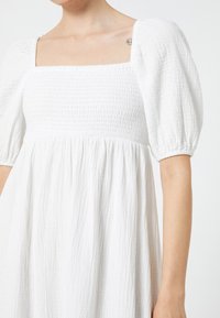 Vestido blanco con escote cuadrado, cuerpo fruncido y mangas abullonadas. Fabricado en tejido texturizado con un corte fluido y holgado.