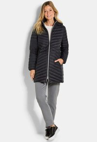 Vrouw in een zwarte, gewatteerde, knielange jas, grijze jeans en zwarte platform sneakers, staande met één hand in de zak, lachend.