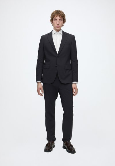 Homme debout portant un costume noir ajusté avec une chemise blanche à col et des chaussures habillées marron foncé sur un fond blanc uni.