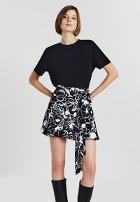 Femme portant un haut noir à manches courtes, une mini-jupe florale noir et blanc avec un détail noué, et des bottes noires jusqu'aux genoux, se tenant avec les mains sur les hanches.