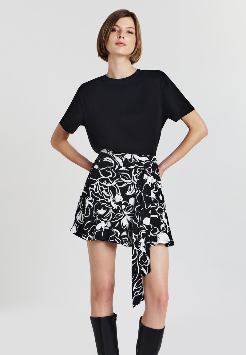 Femme portant un haut noir à manches courtes, une mini-jupe florale noir et blanc avec un détail noué, et des bottes noires jusqu'aux genoux, se tenant avec les mains sur les hanches.