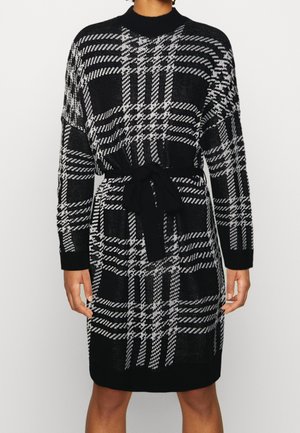 Robe en maille à carreaux noir et blanc, manches longues, col montant, avec ceinture noire à nouer à la taille, portée à mi-cuisse.