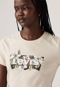 Beżowy bawełniany t-shirt z kwiatowym wzorem, na którym znajdują się białe kwiaty i zielone liście, wyróżniający się logo "Levi's" w wyraźnych białych literach.