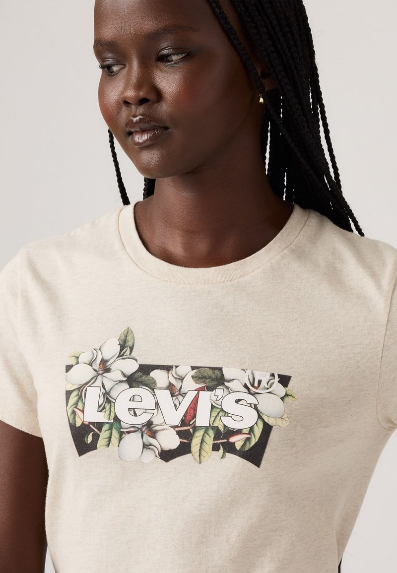 T-shirt en coton beige présentant un motif floral avec des fleurs blanches et des feuilles vertes, mis en valeur par le logo "Levi's" en lettres blanches en gras.