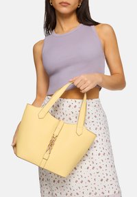 Gul läder tote-väska med två korta handtag, texturerad yta och en guldkedjeaccent på framsidan. Modellen håller väskan.