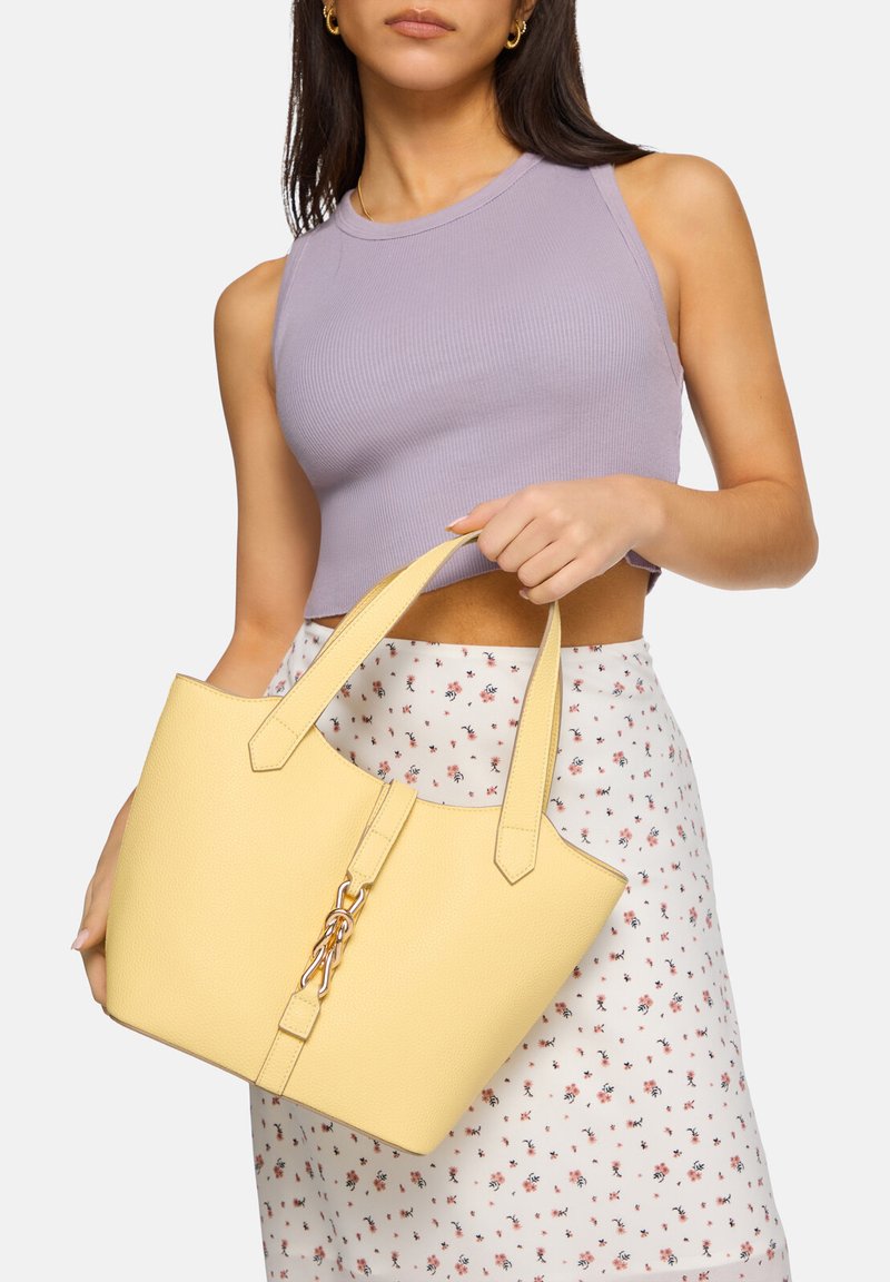 Gul läder tote-väska med två korta handtag, texturerad yta och en guldkedjeaccent på framsidan. Modellen håller väskan.