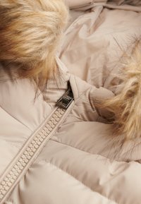 Chaqueta acolchada beige con una textura suave, que presenta una cremallera destacada y detalles de pelo sintético en la capucha, resaltando su diseño acolchado.