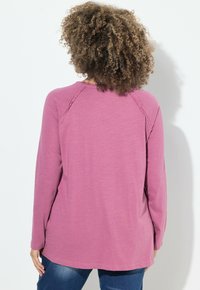 Maglia a maniche lunghe rosa heather con scollo rotondo, dotata di maniche raglan e orlo posteriore leggermente più lungo, indossata con jeans blu.