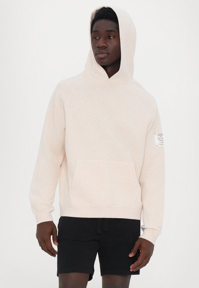 Light beige hooded sweatshirt with a front pocket, textured fabric, and a tag on the right sleeve. Paired with black shorts.