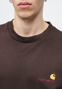 Brown cotton t-shirt with a crew neckline, featuring a small yellow and red Carhartt logo on the left chest area. Smooth texture.