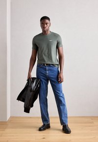 Tricou verde măsliniu cu mânecă scurtă, blugi drepți din denim albastru, geacă din piele neagră aruncată pe un braț, cizme negre până la gleznă, fundal cu podea din lemn.