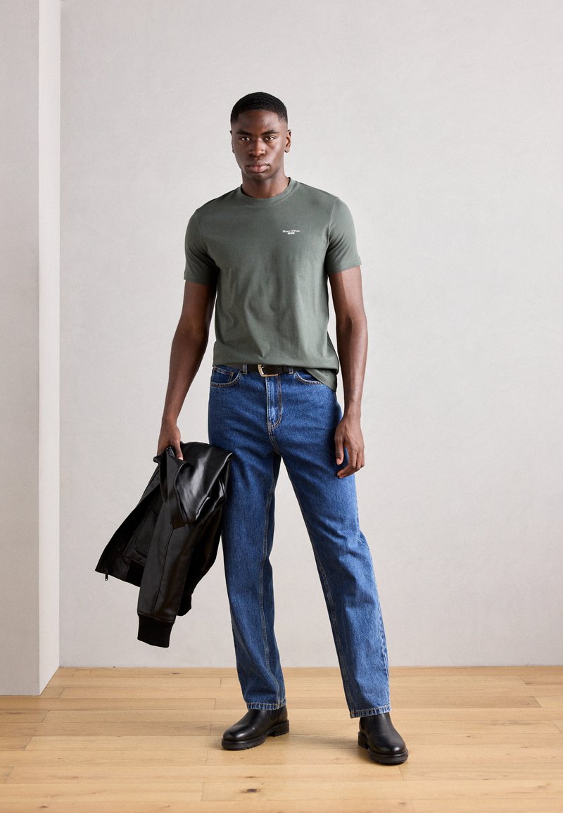 Tricou verde măsliniu cu mânecă scurtă, blugi drepți din denim albastru, geacă din piele neagră aruncată pe un braț, cizme negre până la gleznă, fundal cu podea din lemn.