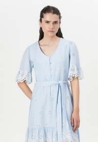Abito azzurro chiaro con maniche corte a campana, motivi floreali ricamati, chiusura a bottoni frontale e cintura legata in vita. Materiale in cotone con orlo arricciato.