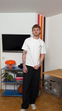 Νεαρός άνδρας με τατουάζ, ντυμένος με λευκό μπλουζάκι και μαύρο παντελόνι, στέκεται σε δωμάτιο με ένα καλάθι μπάσκετ, πορτοκαλί λάμπα και τηλεόραση στον τοίχο.