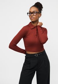 Μπλούζα πόλο σε κοντή γραμμή με μακριά μανίκια σε χρώμα σκουριάς και άνοιγμα με κουμπιά. Συνδυάζεται με ψηλόμεσο μαύρο παντελόνι με ζώνη και διακοσμητικές θηλιές.