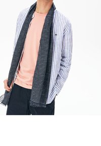 Chemise rayée bleu et blanc portée sur un t-shirt pêche, associée à une écharpe noire à carreaux. Un pantalon bleu marine à fines rayures complète le look.