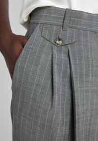 Harmaat raidoitetut housut, joissa on laskoslovi, sivutasku ja nappidetail. Kangas on sileäpintainen ja istuvuus on hyvin muotoiltu.