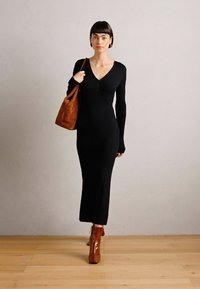 Vestido negro de punto acanalado con escote en V, mangas largas y silueta ajustada, combinado con un bolso de cuero marrón y botas de tacón marrones.