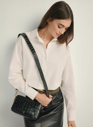 Kobieta w białej koszuli zapinanej na guziki i czarnej skórzanej spódnicy trzymająca czarną, teksturowaną torebkę na ramię z szerokim paskiem.