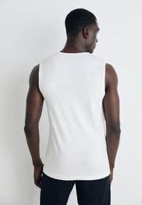 White sleeveless tank top in a smooth cotton fabric, featuring a round neckline and a basic fit, worn with dark pants.