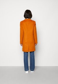 Abrigo largo naranja con un diseño estructurado, que presenta un cuello de muesca, corte recto y abertura en la parte posterior. Combinado con vaqueros azules y zapatillas blancas.