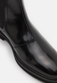 Botă din piele neagră cu un finisaj lucios, panou lateral elastic și detalii cusute. Talpa este robustă, cu o margine texturată.