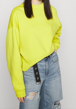Personne portant un sweat-shirt jaune vif à manches longues et un jean bleu clair déchiré avec une ceinture noire pendante.