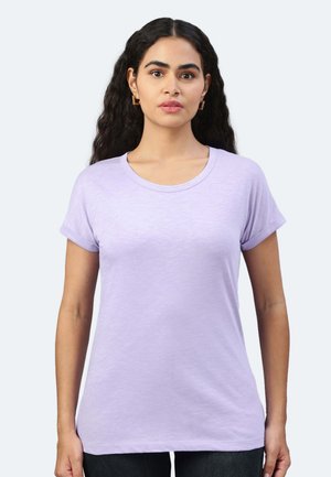 Donna con lunghi capelli ricci che indossa una maglietta semplice viola chiaro a maniche corte e orecchini quadrati dorati, rivolta frontalmente su uno sfondo neutro.