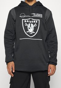 Černá mikina s hladkým materiálem má velké logo Raiders, bílé akcenty a pruhovaný design na ramenou. Dlouhé rukávy.