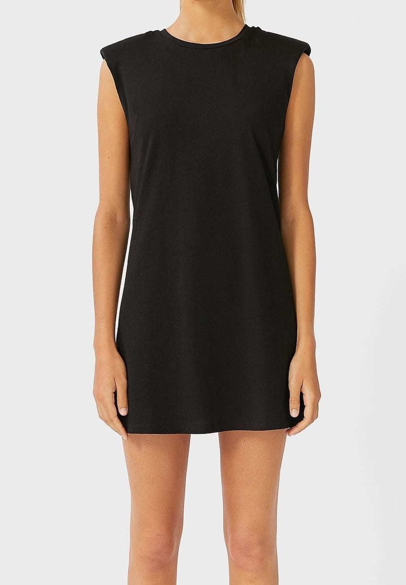 Femme portant une mini-robe noire sans manches avec des épaules rembourrées, se tenant debout, les bras détendus le long du corps devant un fond uni.