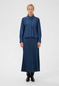 Farmerruha hegyes gallérral, mellzsebbel, párosítva egy plisszírozott, bokáig érő sötétkék szoknyával és fekete térd fölé érő csizmával.