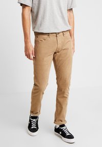 Béžové kordové kalhoty slim fit s jemnou texturou, předními kapsami a zipem, doplněné o černé tenisky.