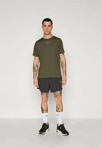 Olivenfarvet T-shirt med HYROX-logo, parret med sorte shorts. Sorte træningssko og hvide sokker fuldender outfittet. Tatoveringer er synlige på armene.