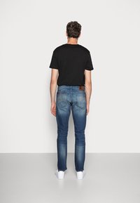 Tmavě šedé tričko s krátkými rukávy v kombinaci se středně modrými džíny. Džíny mají jemné vyblednutí a hnědou koženou nášivku na zadní straně. Bílé tenisky.