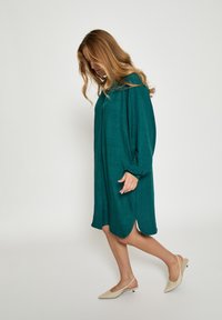 Velika obleka v barvi zeleno-modre z teksturirano tkanino, dolgimi rokavi, stranskimi razporki in sproščenim krojem, kombinirana z naglavnimi čevlji gola barve z ostro konico.