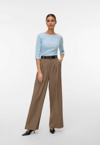 Vrouw staand met hand in de zak, gekleed in een lichtblauw driekwart mouw shirt, bruine wijde broek met zwarte riem, en zwarte spitse hakken.