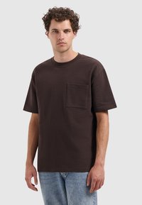 Donkerbruine korte mouwen t-shirt met een ronde hals en een borstzak. De stof heeft een gestructureerde afwerking, gecombineerd met lichtblauwe jeans.