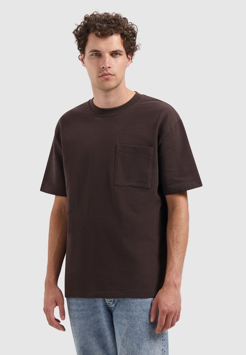 Donkerbruine korte mouwen t-shirt met een ronde hals en een borstzak. De stof heeft een gestructureerde afwerking, gecombineerd met lichtblauwe jeans.