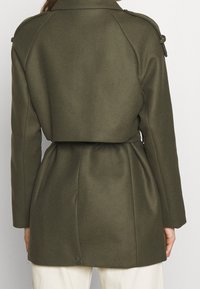 Abrigo de mezcla de lana en verde oliva con cintura ceñida, hombros estructurados y detalles de pliegue en la parte posterior. Textura suave y corte hasta la cadera.