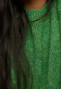 Grüner gestrickter Pullover mit einer strukturierten, gerippten Struktur. Das Material ist weich und weist verschiedene Grüntöne auf.