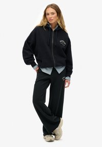Vrouw staat in een zwarte zip-up hoodie en bijpassende wijde broek, met beige sneakers en een gestreept shirt dat eronderuit steekt.