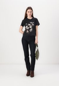 T-shirt nera in cotone con stampa floreale, jeans in denim scuro e stivali alla caviglia marroni. Il modello tiene una borsa verde. Design semplice e moderno.