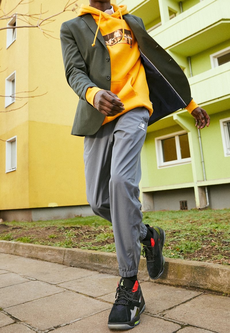 Personne marchant en extérieur portant un sweat à capuche jaune, un blazer sombre, un pantalon de jogging gris et des baskets noires près de bâtiments verts et jaunes.