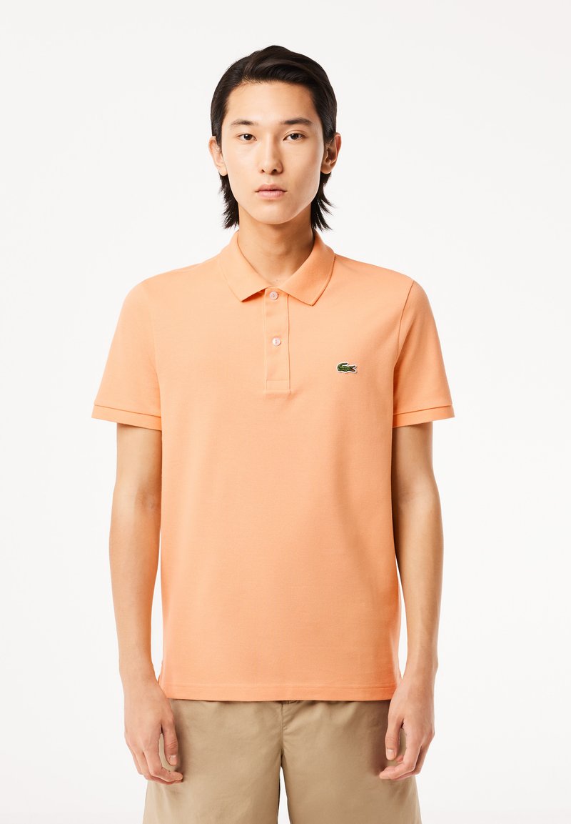 Camisa polo de algodón naranja con cuello clásico y tapeta de tres botones. Logo bordado en verde en el lado izquierdo del pecho. Mangas cortas.