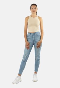 Frau steht entspannt mit hängenden Händen, trägt ein beiges ärmelloses Crop-Top, hellblaue Jeans, weiße Turnschuhe und einen einzelnen hängenden Ohrring.