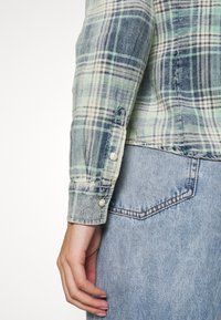 Chemise en flanelle à motif écossais vert, bleu et crème. Possède des poignets boutonnés, une texture douce et un léger délavage. Portée avec un jean.