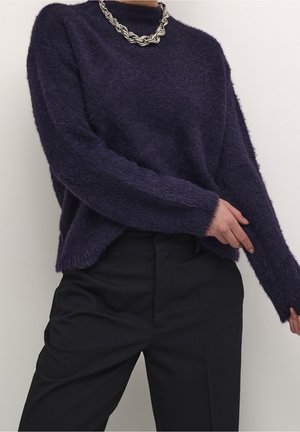 Personne portant un pull duveteux violet foncé, un pantalon noir taille haute et un collier chaîne argenté épais sur un fond uni.